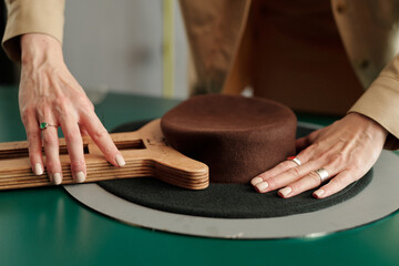 Hands of young female designer forming shape of brown felt porkpie hat with wooden tool by workplace in studio or small craft shop
