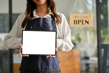 Female barista or waitress in apron stands at the entrance door of her cafe, holding a digital tablet