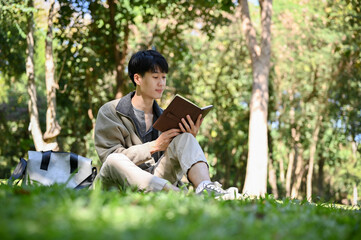 Chilling and focused young Asian male college student reading a book in the park.