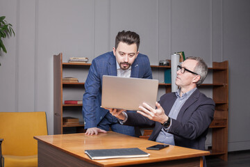 Mature businessman in blue suit is showing work on laptop to his colleague in office