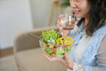 Charming Asian 60s aged woman sipping water and enjoys eating healthy salad bowl