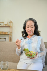 Healthy Asian aged woman enjoys eating salad vegetables mix on sofa in her living room.