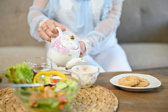 Charming Asian aged woman pouring hot tea from beautiful vintage teapot into a cup