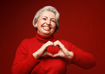 Senior grey-haired woman wearing red sweater smiling in love doing heart symbol shape with hands. - obrazy, fototapety, plakaty