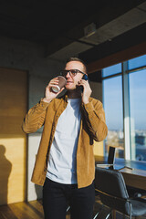 Smiling male worker in casual clothes looking away while talking on mobile phone and drinking coffee during break in modern creative workspace