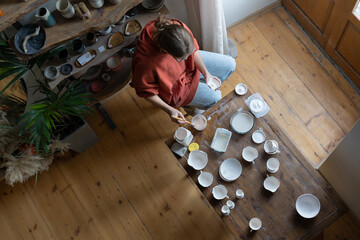 Professional girl potter for home decor shop sits at workplace table, paints finished ceramic products before being fired in kiln. Relaxed young woman making clay plates. Interests for mental health.