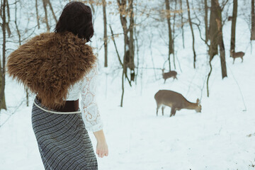 Winter fairytale aesthetic scenic photography. Picture of woman with fur shoulder wrap looking at deer on background. High quality wallpaper. Photo concept for ads, travel blog, magazine, article