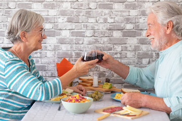 Smiling caucasian senior couple toasting with red wineglass while sitting face to face at table having brunch together at home