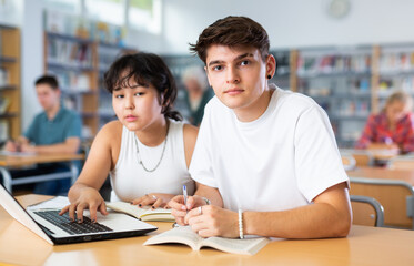 Asian schoolgirl, studying with a classmate guy in the school library on a laptop and making notes in a copybook, preparing for lessons