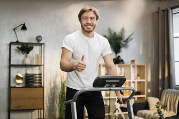 Fototapeta na wymiar Caucasian muscular sporty man in white t-shirt leaned his hands on treadmill while working out in morning at living room at home, looking at camera showing thumb up.