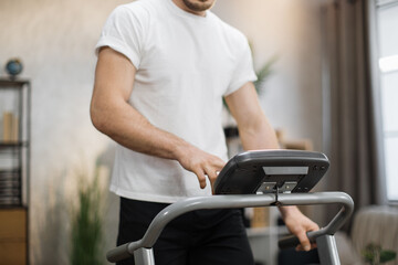 Obraz premium Cropped view of muscular male hands tapping on screen of control panel of treadmill, choosing program of training on background of modern light room.