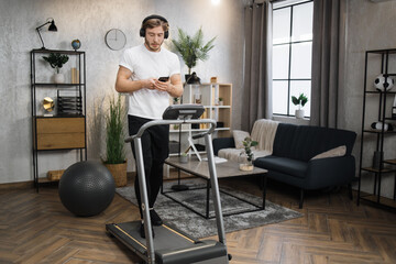 Pozitive caucasian man in sports clothes and sneakers listening music in headphones while using smartphone, doing running exercise on electric treadmill at morning in fitness room at home.