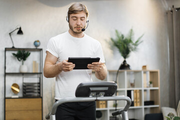 Portrait of active and dynamic young handsome businessman with headset and tablet doing sport fitness at home running on treadmill indoor at night. Tone your body. Perfect shape. Improving endurance.