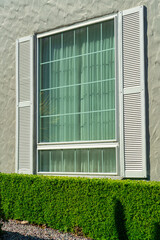 Single window with white shutters and beige stucco exterior walls with front yard shrubs in urban or suburban city