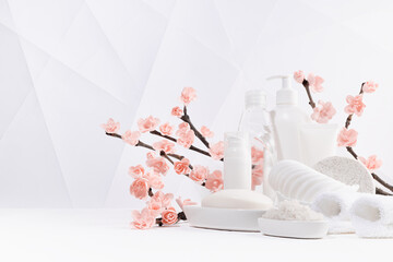 Beauty products for body, skin care in white bottles, branch of spring pink sakura flowers, bath toiletry on table in modern soft light white interior in minimalist geometric japan style, copy space.