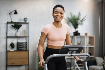 Smiling african american sporty woman in pink t-shirt leaned her hands on treadmill while working...