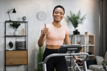 Smiling african american sporty woman in pink t-shirt leaned her hands on treadmill while working out in morning at living room at home, looking at camera showing thumb up.