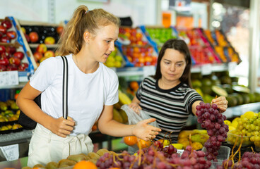 Two positive woman choosing sweet fresh grape at grocery section of supermarket
