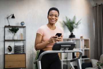 Fototapeta na wymiar Close up view of young sporty african woman using running machine simulator while making selfie or having video call. Healthy sportswoman having workout on treadmill indoors her home.