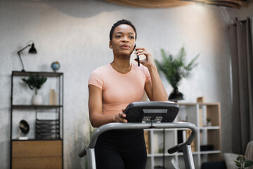 Fototapeta na wymiar Portrait of young african sportive woman in sportswear talking on smartphone while training at home, doing cardio exercise on treadmill. Concept of sport, health care, communication.