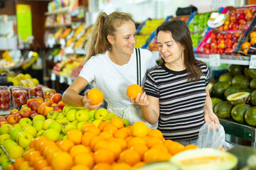 Young woman with a fifteen-year-old girl who came to the supermarket for shopping, choose oranges at the counter, putting ..them in a package