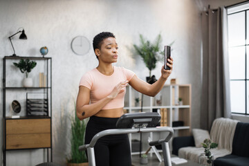 Fototapeta premium Attractive young sports african woman having online video call while working out, running, doing cardio training on treadmill in morning time, indoor on background of modern apartment or gym.