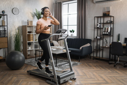 Pozitive african woman in sports clothes and sneakers listening music in headphones while using smartphone, doing running exercise on electric treadmill at morning in fitness room at home.
