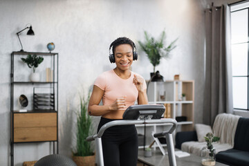 Fototapeta na wymiar Smiling attractive young sports african woman doing fitness exercise, running on treadmill and listening music. Athletic and muscular female having actively workout indoor, at modern apartment.