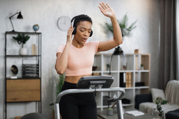 Beautiful african american women with short curly hair in sports clothes and sneakers, listening music in headphones doing running exercise on electric treadmill in fitness room at home.