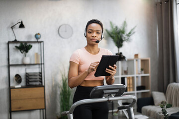 Portrait of active and dynamic young pretty businesswoman with headset and tablet doing sport fitness at home running on treadmill indoor at night. Tone your body. Perfect shape.