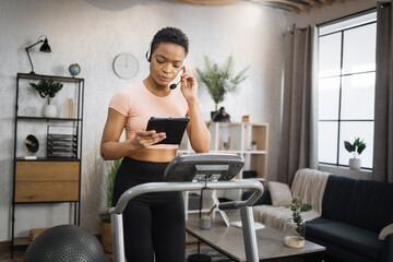 Confident muscular sporty businesswoman in headset working remote from home using digital tablet, while having cardio workout, running on treadmill.