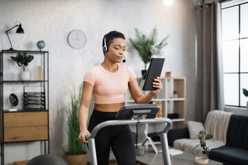 Close up of young active african businesswoman in sportswear and headset training at home working with tablet, doing cardio exercise on treadmill. Concept of sport, business.