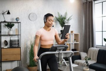 Fototapeta premium Close up of young active african businesswoman in sportswear and headset training at home working with tablet, doing cardio exercise on treadmill. Concept of sport, business.