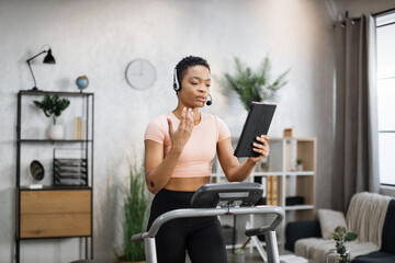 Close up of young active african businesswoman in sportswear and headset training at home working with tablet, doing cardio exercise on treadmill. Concept of sport, business.