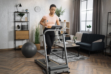 Fototapeta premium Beautiful african woman in sports clothes and headphones holding bottle of water doing cardio training on treadmill at home gym. Concept of sport, health care, action, remote leisure.