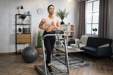 Portrait of active and dynamic young charming thirsty african woman holding water bottle doing sport fitness at home running on treadmill indoor. Tone your body. Perfect shape. Improving endurance.