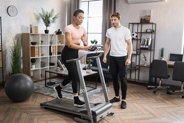 Fototapeta premium Portrait of focused young african female wearing sportswear and using treadmill and her trainer bearded caucasian male. Home fitness workout sporty woman and man training on running track.