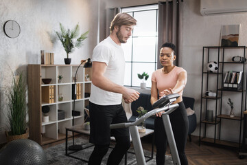 Fit african woman coach using control panel while training young caucasian man to run on treadmill. Attractive sporty fit people using running track and choosing mode for sports training.