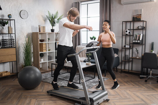 Fit African Woman Coach Using Control Panel While Training Young Caucasian Man To Run On Treadmill. Attractive Sporty Fit People Using Running Track And Choosing Mode For Sports Training.