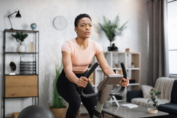Fototapeta na wymiar Portrait of focused young african female athlete wearing sportswear and using exercise bike. Home fitness workout sporty woman training on smart stationary bike indoors.