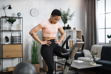 Young attractive sporty tired african american woman in sportswear having morning workout at modern light apartment. Healthy athletic female doing cardio on stationary bike.