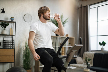 Focused young male wearing sportswear drinking water, using exercise bike. Home fitness workout sporty man training on smart stationary bike indoors. Young caucasian guy athlete.