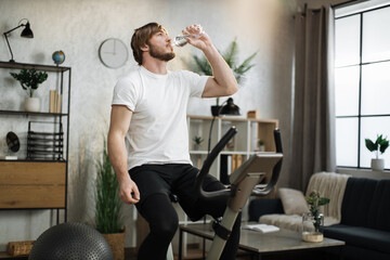 Focused young male wearing sportswear drinking water, using exercise bike. Home fitness workout sporty man training on smart stationary bike indoors. Young caucasian guy athlete.