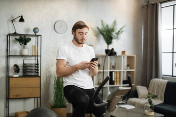 Healthy fit smiling caucasian man training at home on exercise static bike during workout holding phone, watching online lessons for motivation. Male healthy weekly habits app.
