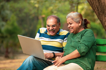 Happy senior couple sitting on bench in the park and using laptop.