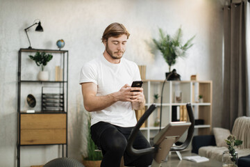 Portrait of focused young male wearing sportswear using exercise bike typing message on phone. Home fitness workout sporty man training on smart stationary bike indoors.