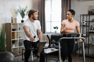 Fototapeta premium Young couple doing cardio on stationary bike and treadmill. Attractive female and male working out at home. Woman running on treadmill and man training on exercise bike.
