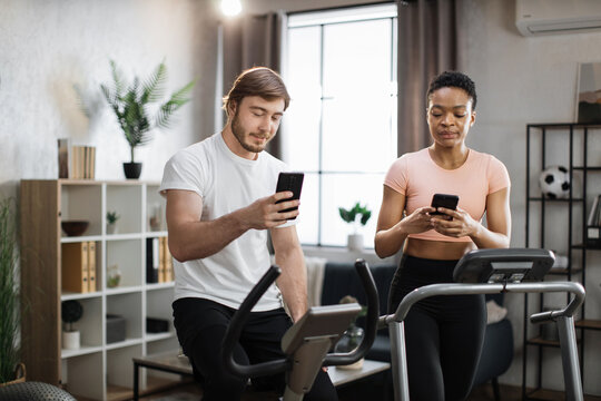 African american sports woman in sportswear running on treadmill and fit caucasian man cycling bike and showing each other photos from social media, networks.
