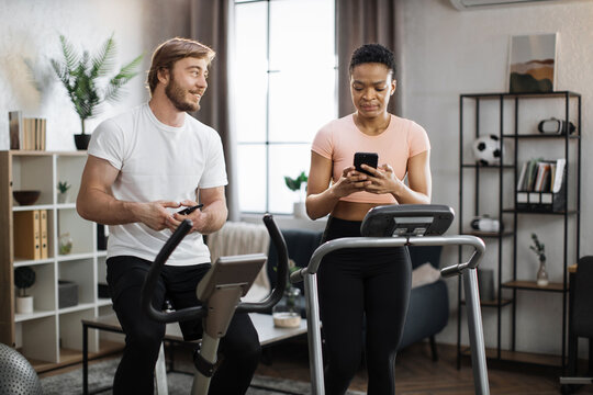 African american sports woman in sportswear running on treadmill and fit caucasian man cycling bike and showing each other photos from social media, networks.