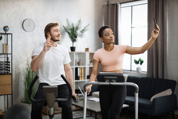 Fototapeta na wymiar African american sports woman in sportswear running on treadmill and fit caucasian man cycling bike and making selfie or having online video call.